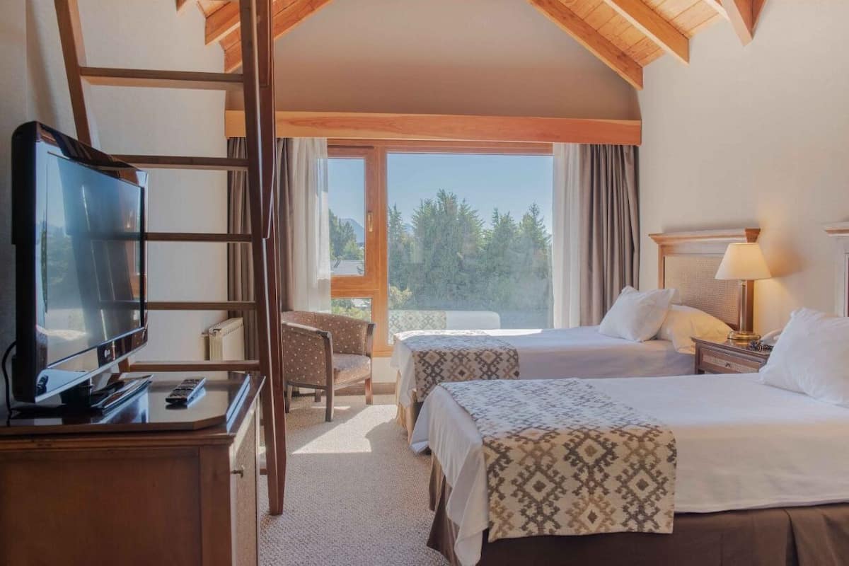 A hotel room with two beds, a TV, a desk, and a chair. There is a large window with curtains and a view of trees.