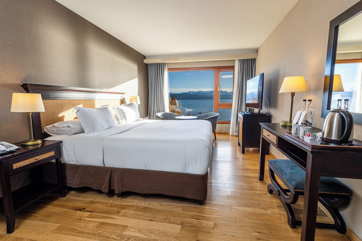 A hotel room with a large bed, a desk, a chair, a TV, and a view of the sea.