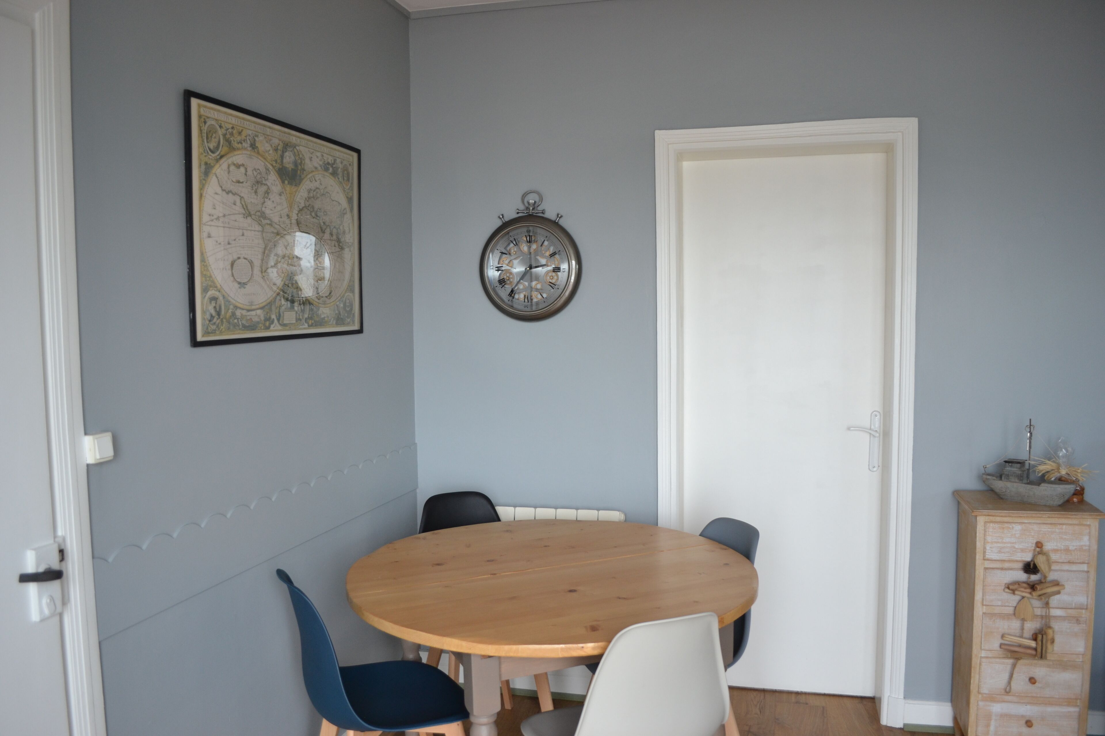 A dining area with a round wooden table, blue and white chairs, a wall clock, a world map, and a wooden cabinet.