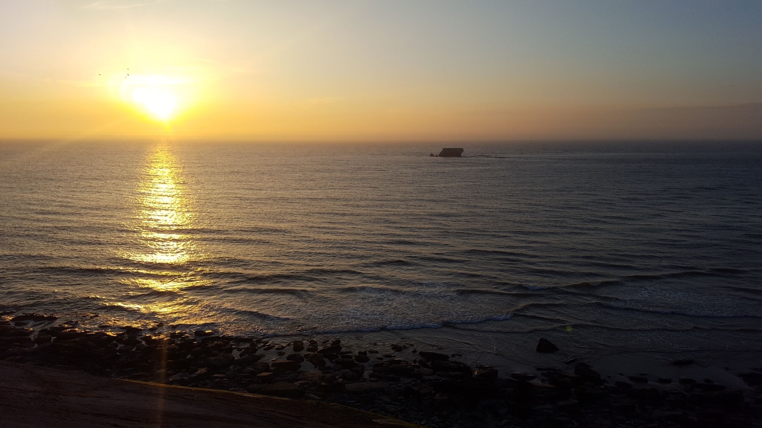 Un coucher de soleil sur une mer calme, avec une côte rocheuse.