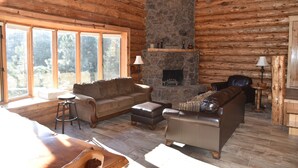 A log cabin interior with a stone fireplace, leather sofas, and large windows offering a view of the surrounding forest. - Log home On the Deschutes River Bend, OR (Bend)