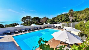 Un espace piscine agrémenté de chaises longues et de parasols, entouré de verdure et offrant une vue sur la mer.