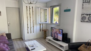 A modern living room with a white sofa, a coffee table with remote control, a TV on a white stand, and a window with a view of greenery. - BEAUTIFUL VILLA WITH HEATED JACUZZI AND WIFI, facing the golf course in Lacanau-Océan; sleeps 6, 3 bedrooms, 2 bathrooms (Lacanau)