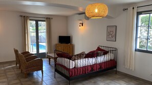 Une chambre avec un lit, une télévision, un fauteuil en osier, une table à manger et une vue sur une piscine.