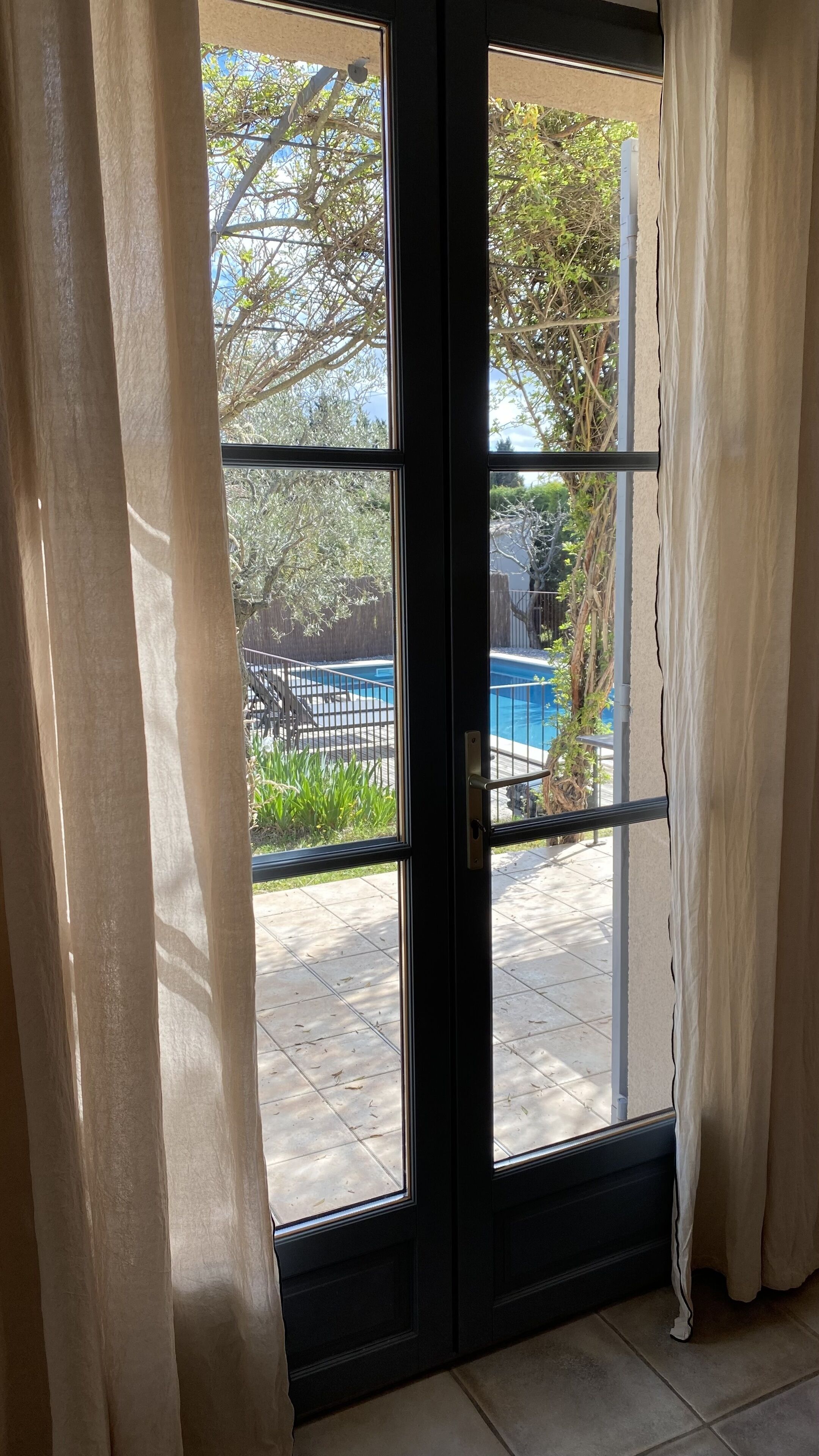 A view through a glass door with a metal handle, leading to a backyard with a swimming pool, a wooden deck, and a metal fence.