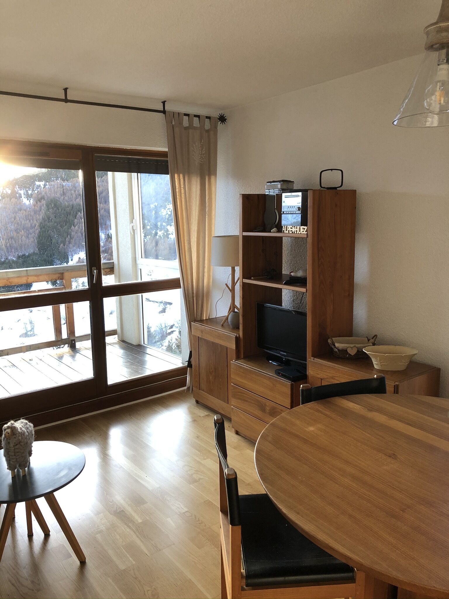 A room with a wooden dining table, a TV on a wooden stand, a lamp, and a view of a snowy landscape through a large window.