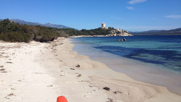 Ein Strand mit klarem Wasser, einem sandigen Ufer und einem Turm auf einer felsigen Insel.