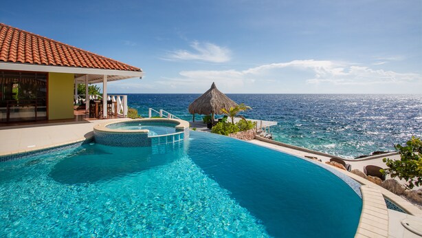 Ein Pool mit gewölbter Kante, ein Gebäude mit Reetdach und Blick auf den Ozean.