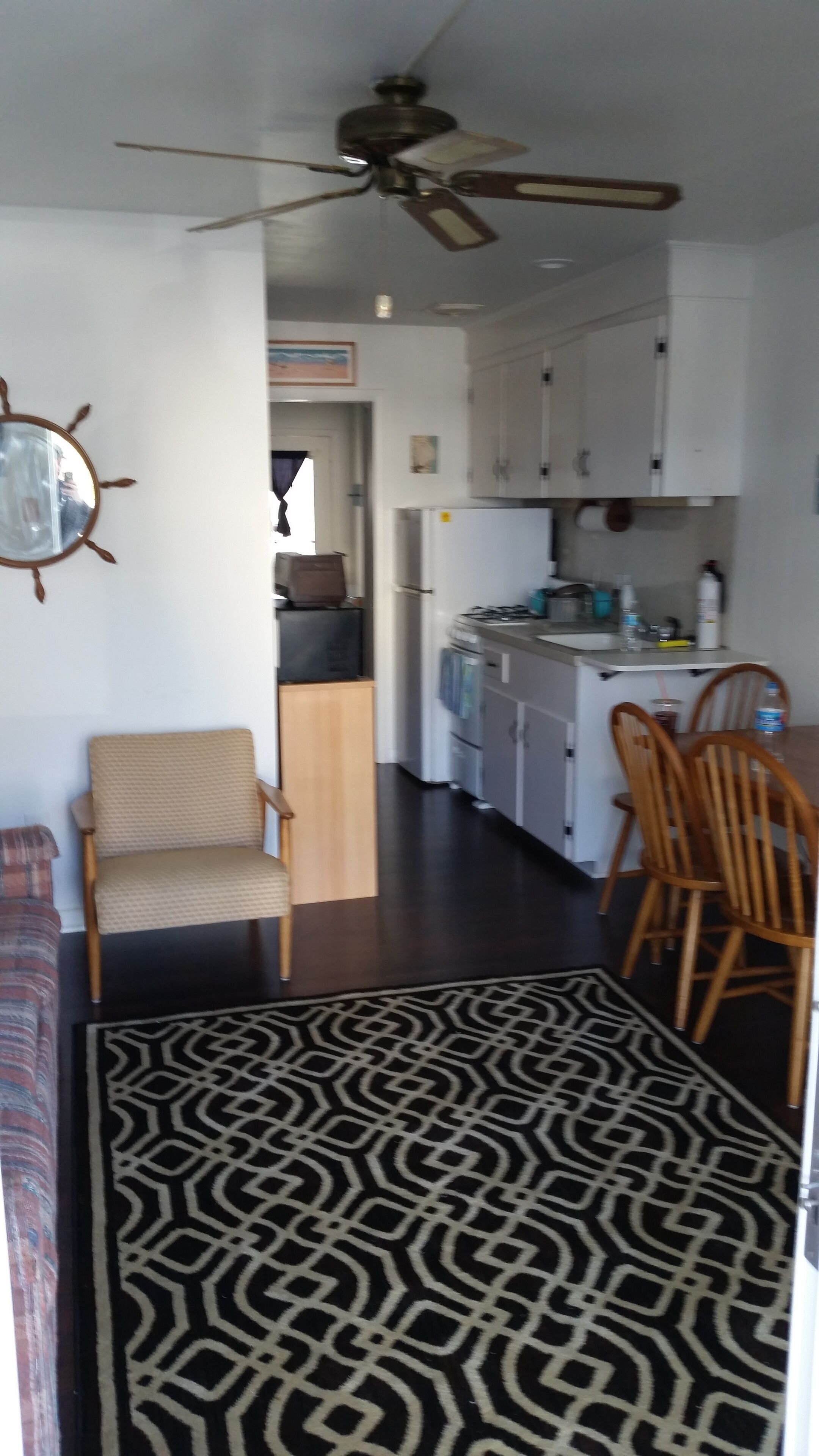 A compact living space with a kitchenette, a dining area with wooden chairs, a patterned rug, and a ceiling fan.