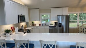 Uma cozinha moderna com armários brancos, eletrodomésticos de aço inoxidável, uma ilha central com banquetas e grandes janelas com vista para o exterior.
