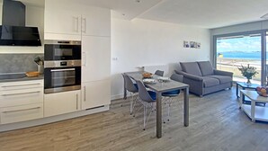 A modern kitchen with white cabinets and stainless steel appliances, a dining area with a table and chairs, and a gray sofa. - Spacious studio with terrace, sea and shops 700m, private parking, Wi-Fi included (Larmor-Plage)