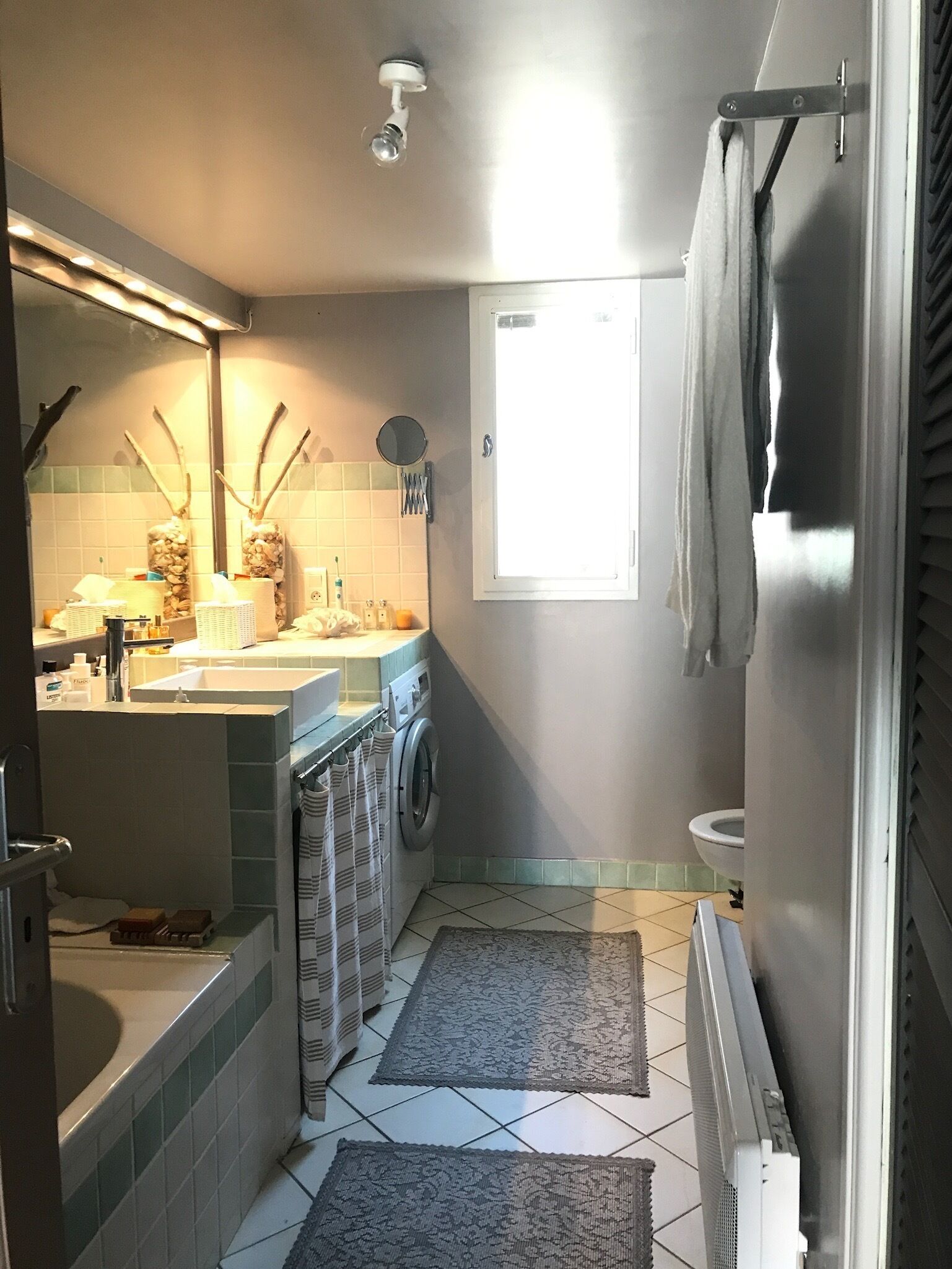 A compact bathroom with a sink, mirror, and washing machine. There is a window providing natural light and a towel rack.