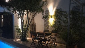An outdoor patio with a pool, wooden table, chairs, and potted plants.