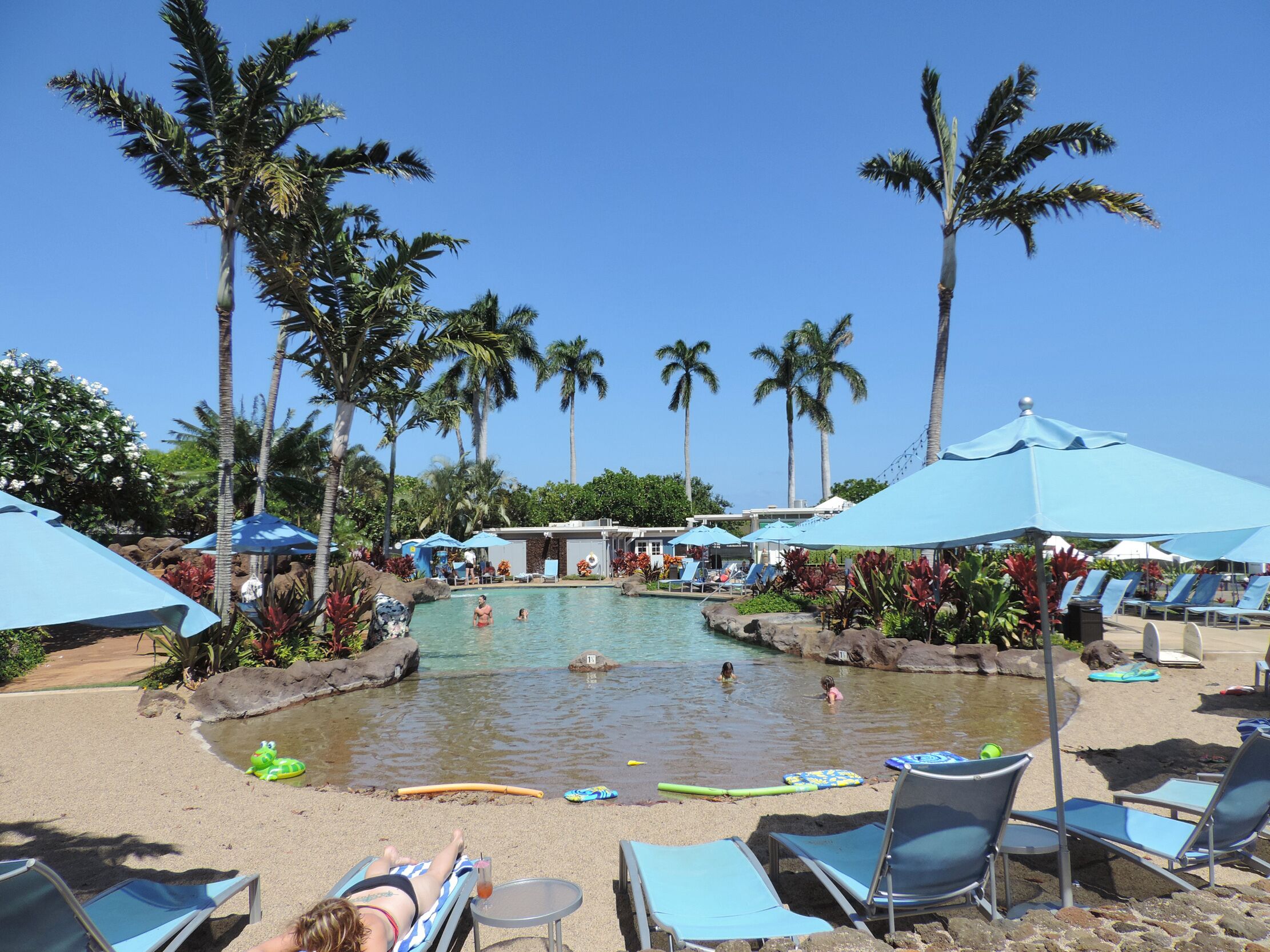 Zona de piscina con sillones de descanso y sombrillas, rodeada de palmeras.