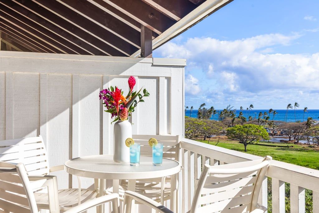 Een wit buiten zitgedeelte met een tafel en stoelen, een vaas met bloemen en uitzicht op de oceaan en palmbomen.