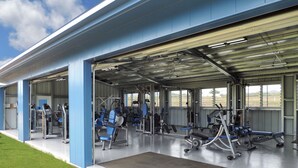 Une salle de sport extérieure abritée, dotée de divers équipements d’exercice, notamment des tapis de course et des appareils de musculation, le tout sous un toit bleu.