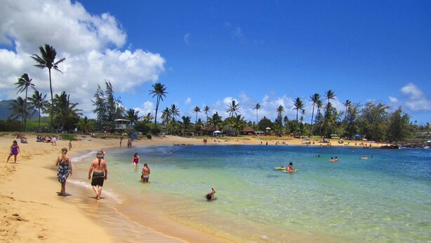 Une plage à l’eau claire, bordée de palmiers, où les gens se détendent sur le sable et se baignent.