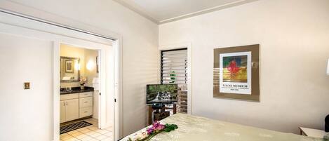 A hotel room with a bed, a TV, a framed picture on the wall, and a bathroom visible through an open door.