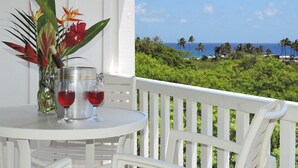 Une table blanche placée à l’extérieur, avec deux verres à vin et un récipient en métal, le tout mis en valeur par un paysage verdoyant luxuriant et l’océan en arrière-plan.