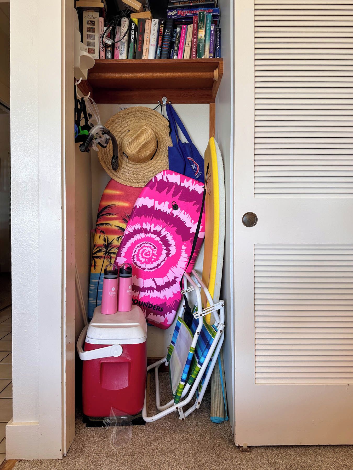 A storage area with a shelf holding books, a straw hat, a pink towel, a red cooler, and a surfboard.