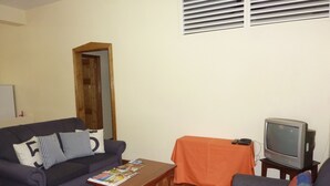 Ein Wohnzimmer mit einer Couch, einem Couchtisch mit Zeitschriften, einem Fernseher auf einem Schrank und einem gemusterten Teppich.
