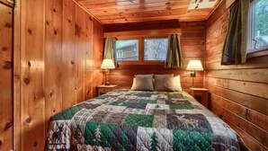A wooden cabin bedroom with a quilted bedspread, two bedside lamps, and a window with curtains.