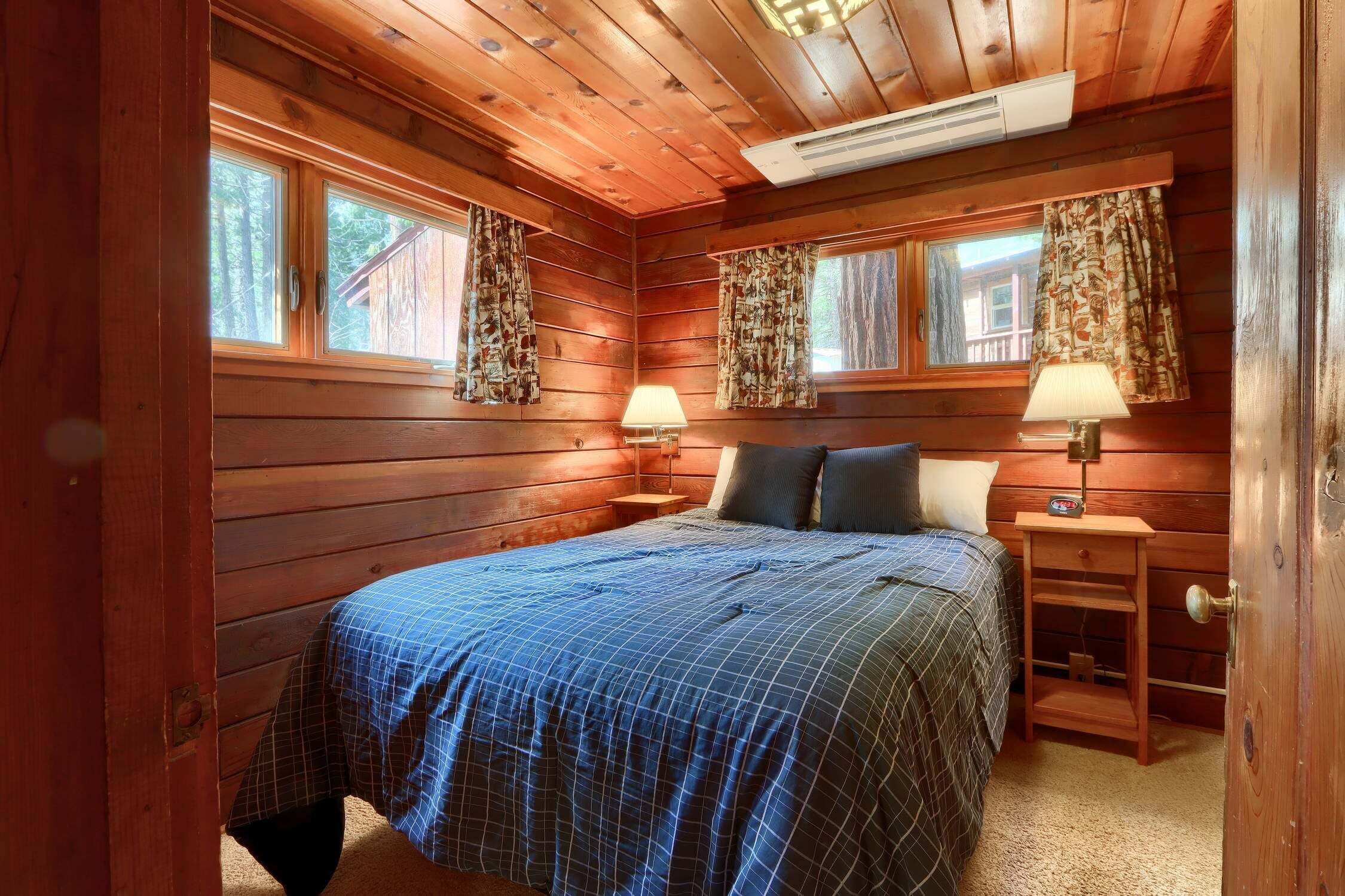 Une chambre dans une cabane en bois, avec un lit, deux tables de chevet équipées de lampes et une fenêtre avec des rideaux.