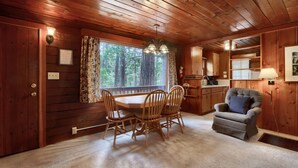 A wooden cabin interior with a dining table, chairs, a sofa, a window with curtains, and a kitchen area.