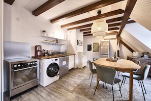 Une cuisine compacte dotée d’appareils électroménagers modernes, un coin repas avec une table et des chaises en bois, et des poutres apparentes en bois au plafond.