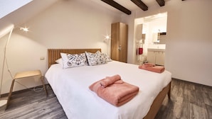 Un dormitorio con cama, cabecera de madera, mesita de noche, lámpara, armario y baño visible a través de una puerta abierta.