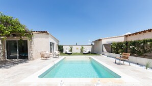 Una piscina con bordo piastrellato, due sdraio e un piccolo giardino con una panchina.