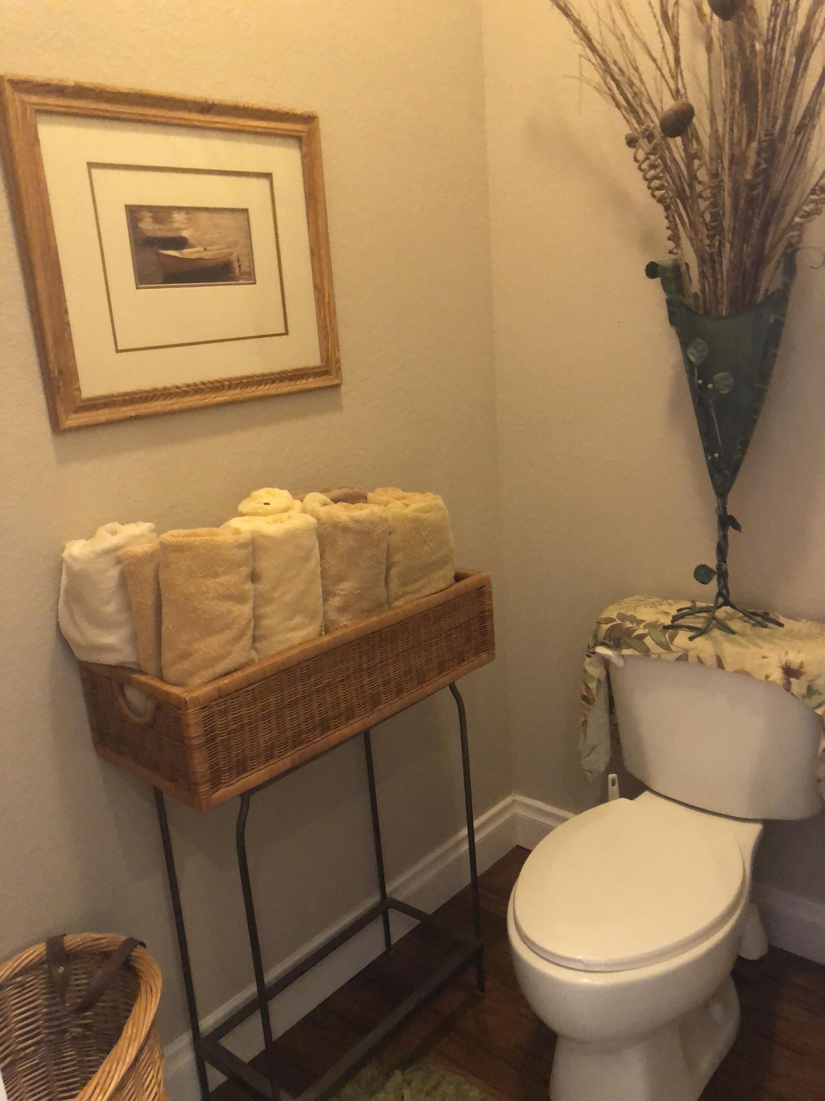Une salle de bain avec un panier en osier contenant des serviettes, un tableau encadré au mur et un vase avec des plantes séchées.