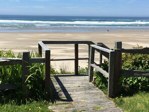 Ein hölzerner Steg führt zu einem Strand, im Hintergrund sind Wellen zu sehen.