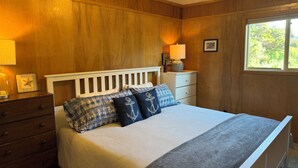 A bedroom with a wooden paneled wall, a white bed with striped bedding, a nightstand with a lamp, and a window with a view of greenery. - Classic Honeymoon Beach Cottage. Private, Romantic, Oceanfront.  Pet friendly. (Waldport)