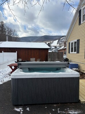 Un jacuzzi en un patio trasero con terraza de madera, cerca una valla blanca y una casa al fondo.