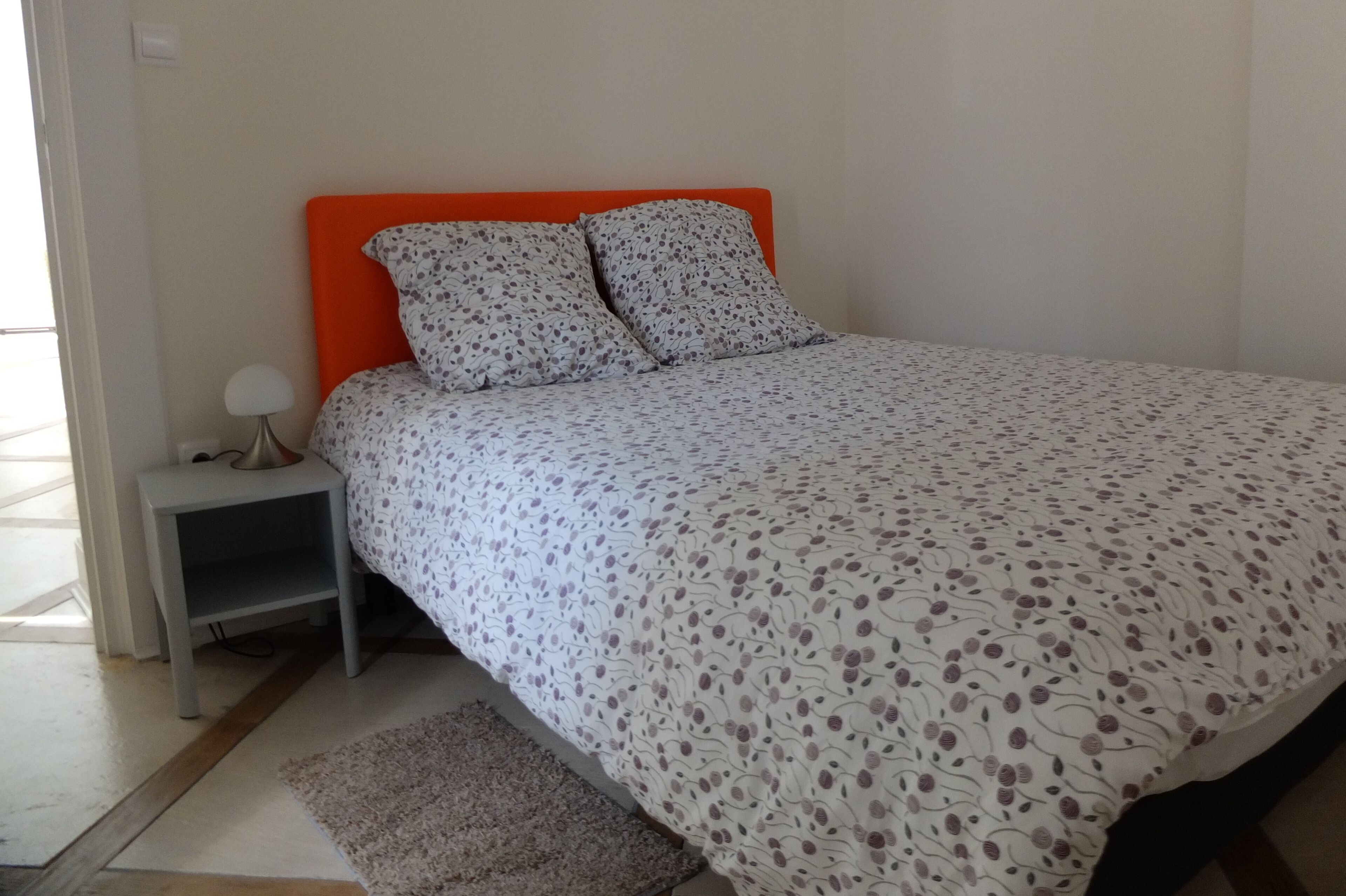 Un dormitorio con cama, cabecera naranja, mesita de noche con lámpara y una alfombra pequeña.