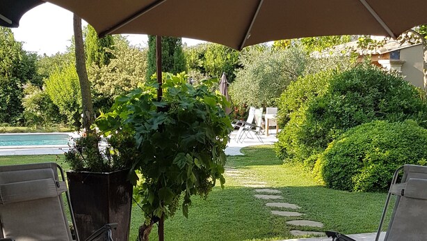 A patio area with two lounge chairs under a large umbrella, surrounded by greenery and a swimming pool in the background.