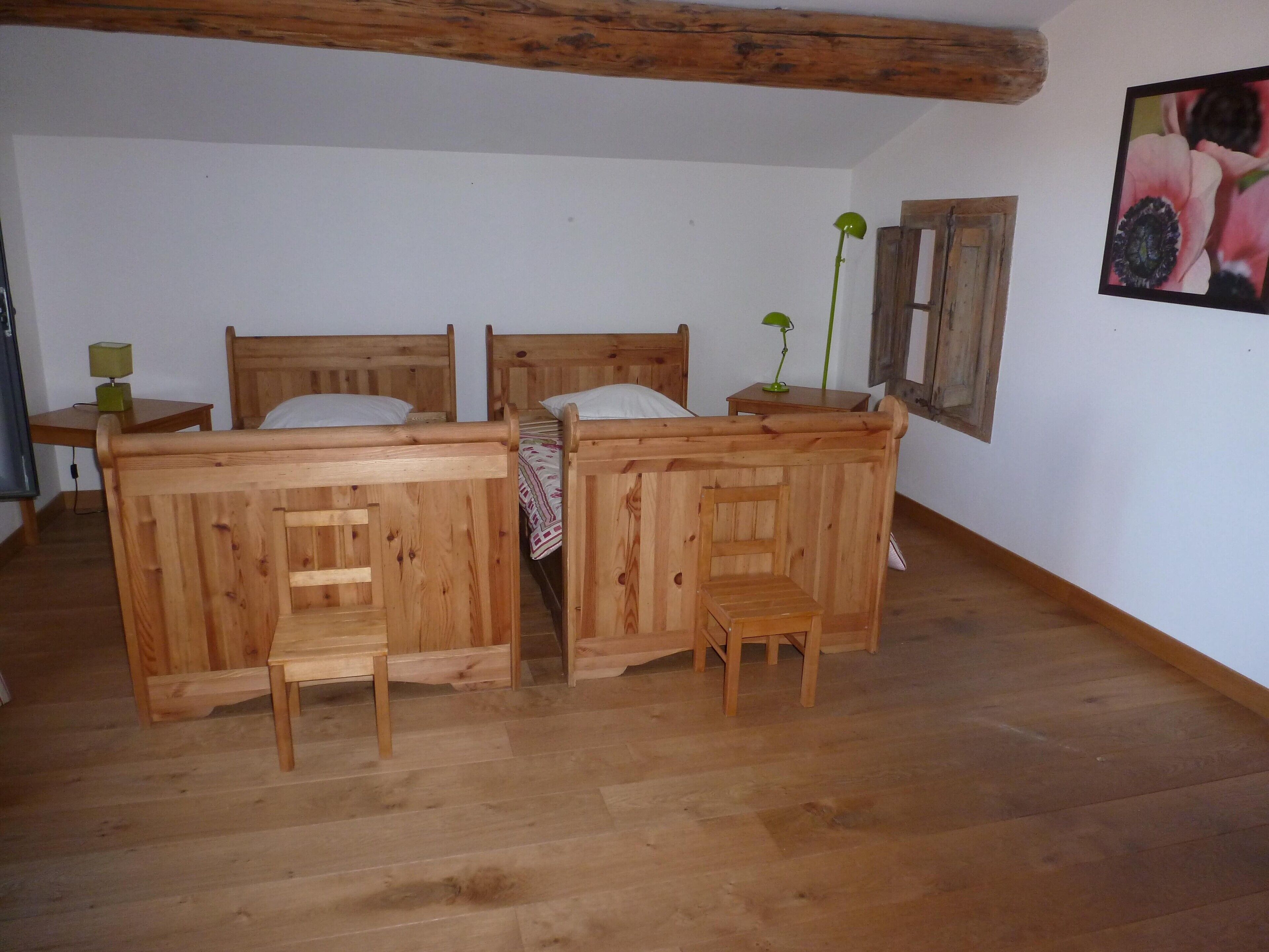 Una stanza con due letti di legno, una piccola sedia in legno, un tavolo con una lampada e un quadro appeso al muro.