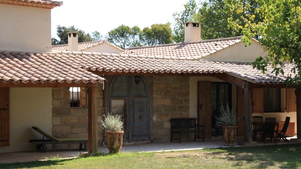 Un bâtiment en pierre avec un toit en tuiles, un banc en bois et une table avec des chaises à l’extérieur.