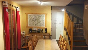 Ein Essbereich mit einem langen Holztisch und Stühlen, eine Küchenzeile mit Schränken und einer Karte an der Wand sowie eine Treppe, die zu einer oberen Ebene führt.
