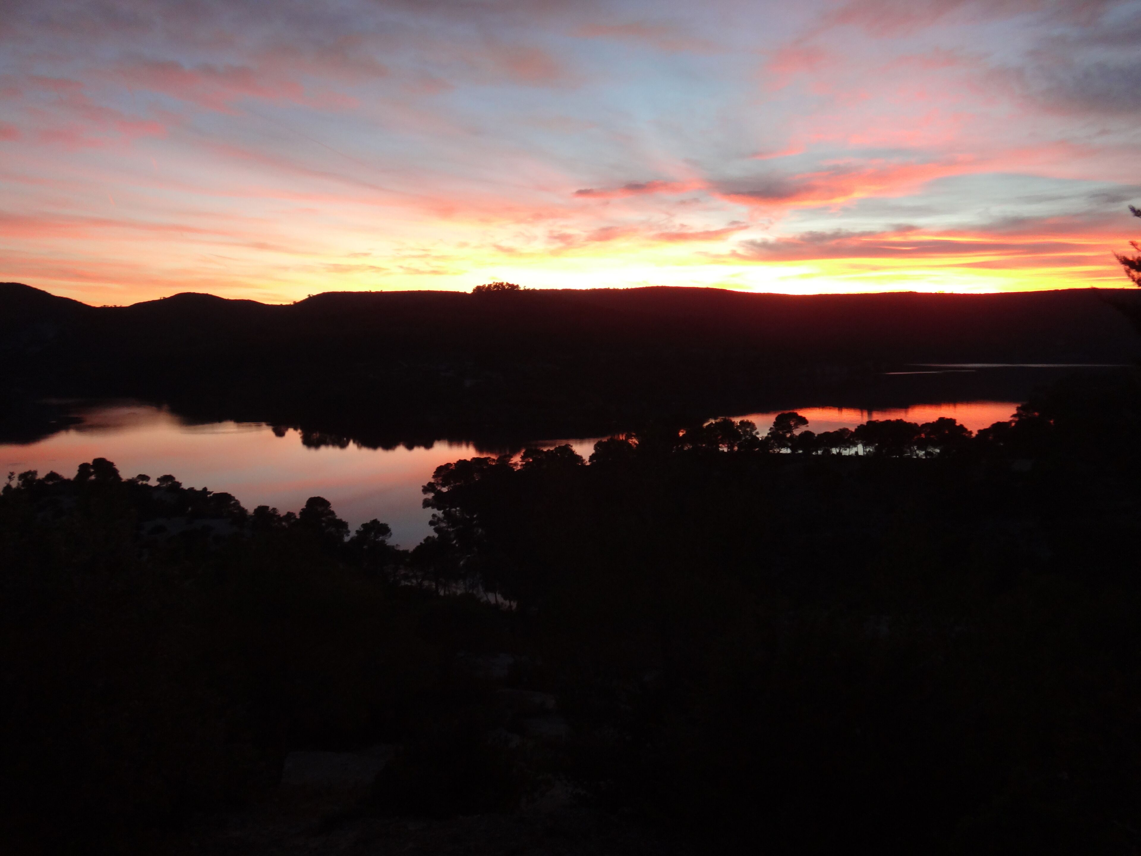En solnedgång över en sjö, med silhuetter av träd och kullar i bakgrunden.