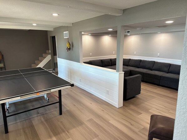 Une pièce avec une table de ping-pong, un canapé d’angle et un escalier.