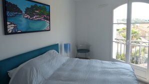 A bedroom with a bed, a painting, a chair, and a window with a view of buildings. - Place of the Strings in the heart of St Tropez apartment T2 new 2/4 with parking (Saint-Tropez)