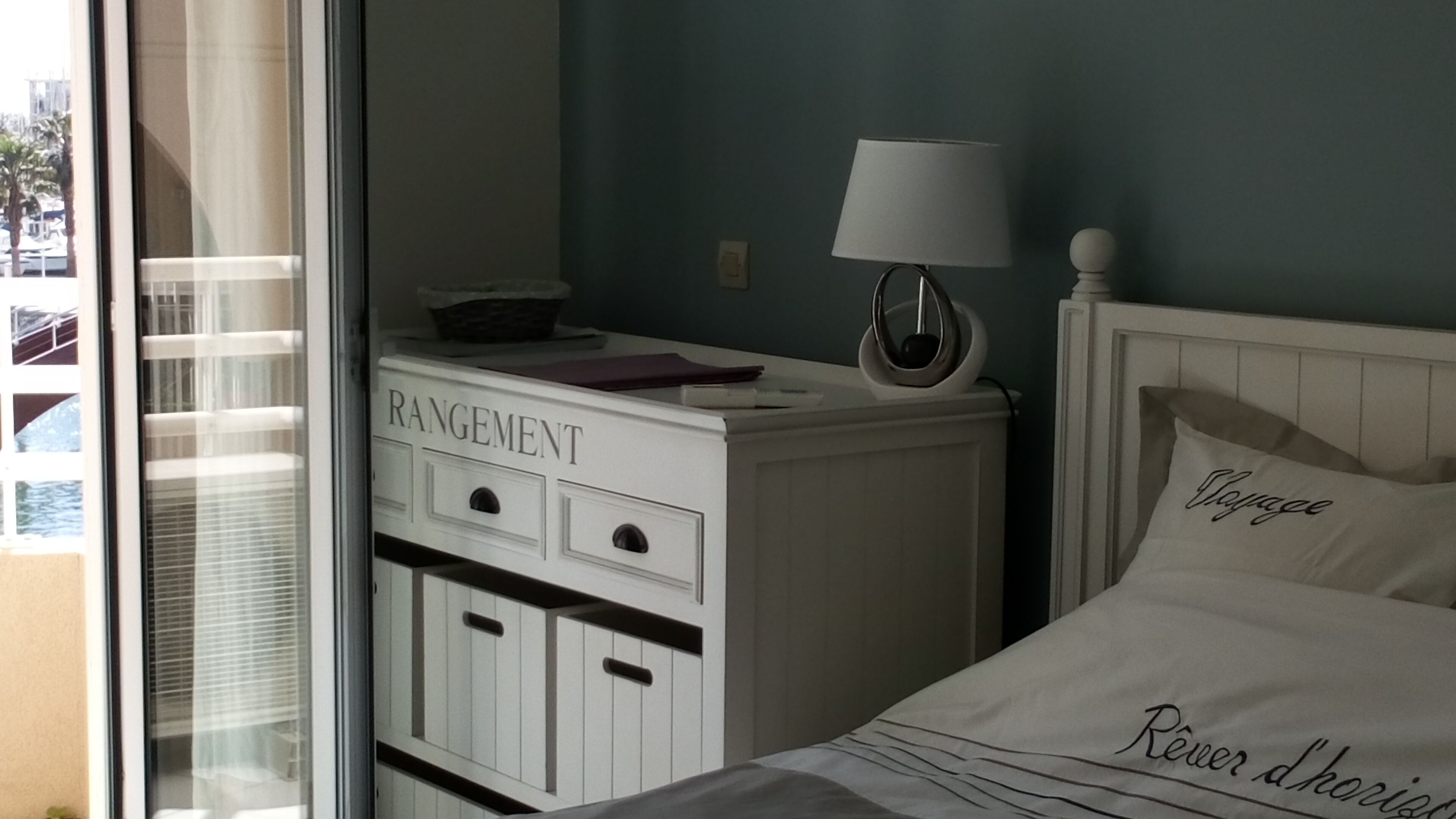A bedroom with a bed, a nightstand, a lamp, a dresser labeled 'RANGEMENT,' and a window with blinds.