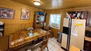 Ein Zimmer mit Holzwandverkleidung, einem Esstisch, Stühlen, einem Schrank, einem Fenster mit Jalousien und einem Kühlschrank.