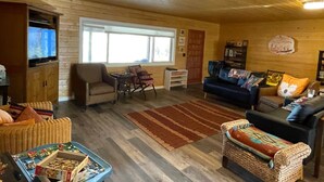 Ein gemütliches Wohnzimmer mit Holzboden, einem Fernseher, einer Couch, einem Sessel, einem kleinen Tisch und einem Bücherregal.