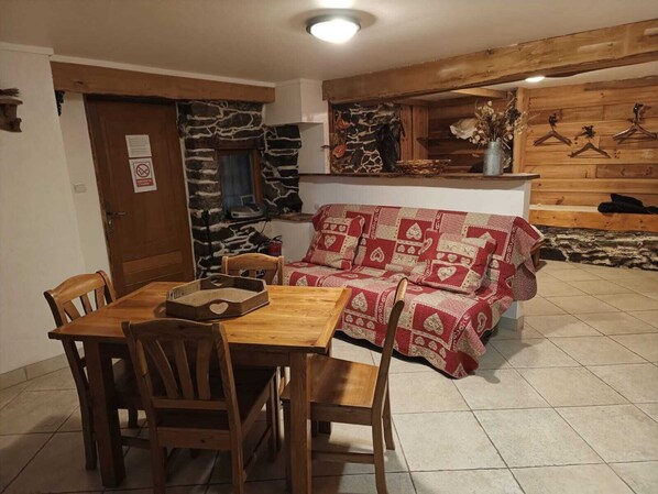Una habitació acollidora amb una taula de dinar i cadires de fusta, una paret de pedra, un sofà amb una manta estampada i prestatges de fusta amb penjalls.