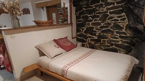 Una cama individual con estructura de madera, ropa de cama color beige y una almohada decorativa roja y blanca. Se ve una pared de piedra a la derecha de la cama.