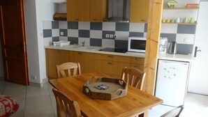 Cocina compacta con armarios de madera, un microondas y un refrigerador. Hay una mesa de comedor con sillas y una bandeja de madera encima.