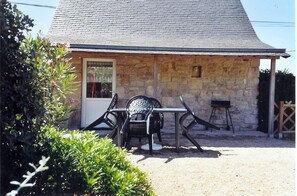 A stone building with a tiled roof, a door, and a small window. There is a table with chairs outside.